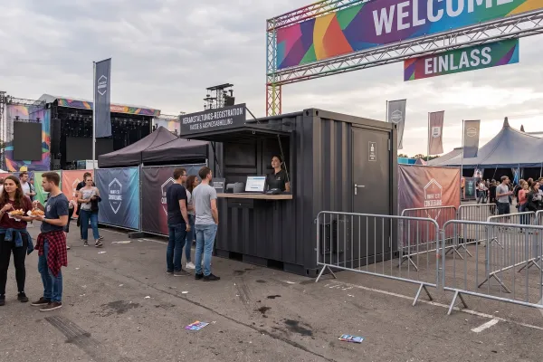 Kompakter Kassen- & Akkreditierungs-Container - Foto Anthrazitfarbener Kassen- und Registrierungscontainer aus Übersee-Bauteilen auf einem Event-Einlassbereich mit geöffneter Verkaufsklappe, Counter-Arbeitsplatz für das Personal und vorgelagerten Absperrgittern unter einem Welcome-Schild.