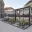 Foto Zweistöckiger, dunkelgrauer Roadshow-Container mit dem Branding "Urban Hydro" auf einem gepflasterten Stadtplatz. Das Erdgeschoss hat bodentiefe Glasfronten und einen vorgelagerten Loungebereich mit Sesseln und Pflanzenkübeln. Eine Außentreppe führt in das verglaste Obergeschoss und auf eine Dachterrasse mit Sonnenschirmen.